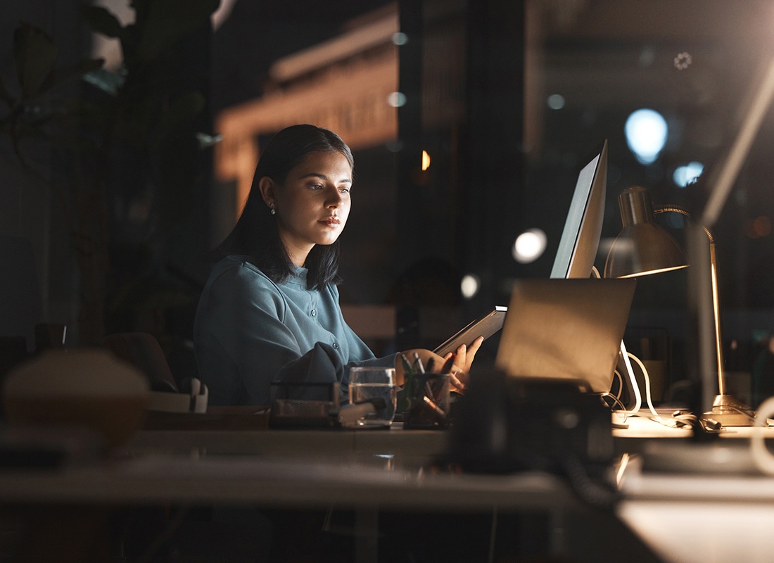 Read Our Reviews - Young Business Woman Sitting in her Office During the Night Working on a Project Using a Tablet and Computer with Scenes of the City Through the Window